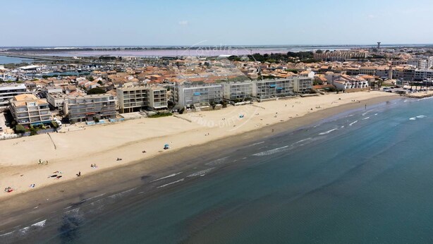 An aerial view of a coastal town with a sandy beach, buildings, and a clear sky.