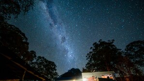 Una casa con un ventanal grande, un espacio exterior cubierto y vistas del cielo nocturno con estrellas y la Vía Láctea.