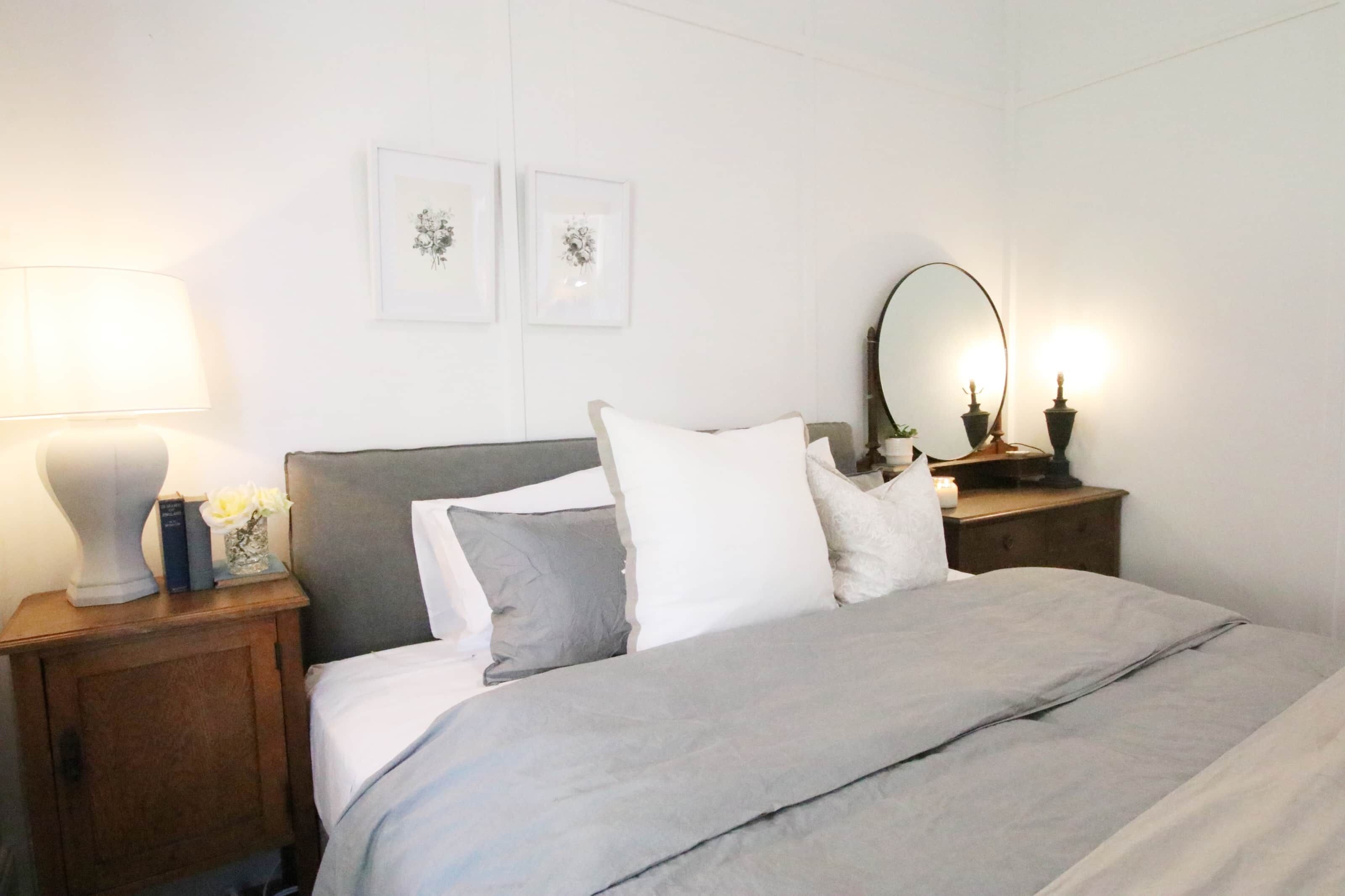Una cama bien hecha con una decoración en tonos grises y blancos, una lámpara de noche, una mesita con un jarrón de flores y dos cuadros en la pared.