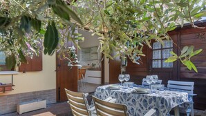 Un área de comedor al aire libre con una mesa lista para dos personas, rodeada de sillas de madera, bajo un árbol.