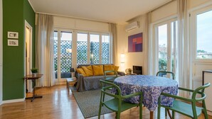Una habitación con sofá, una mesa de comedor con sillas, una mesita con una planta y un ventanal con vista a un balcón.