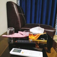 Una habitación con un sillón de cuero marrón, una mesita con un control remoto, una caja de pañuelos, un vaso y una servilleta rosa.