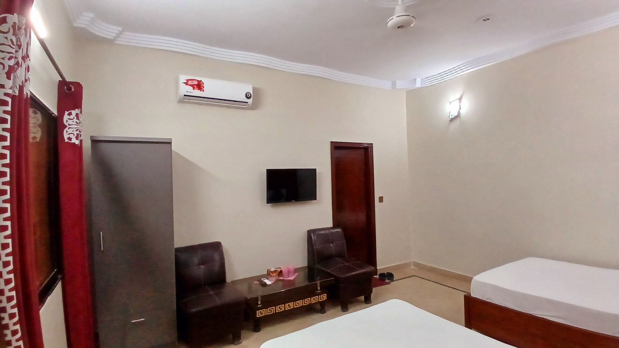 A hotel room with a bed, two chairs, a TV, a wardrobe, and an air conditioning unit.