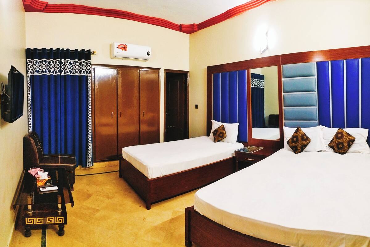 Habitación de hotel con dos camas individuales, una cama grande, un armario, un escritorio con silla y un televisor.