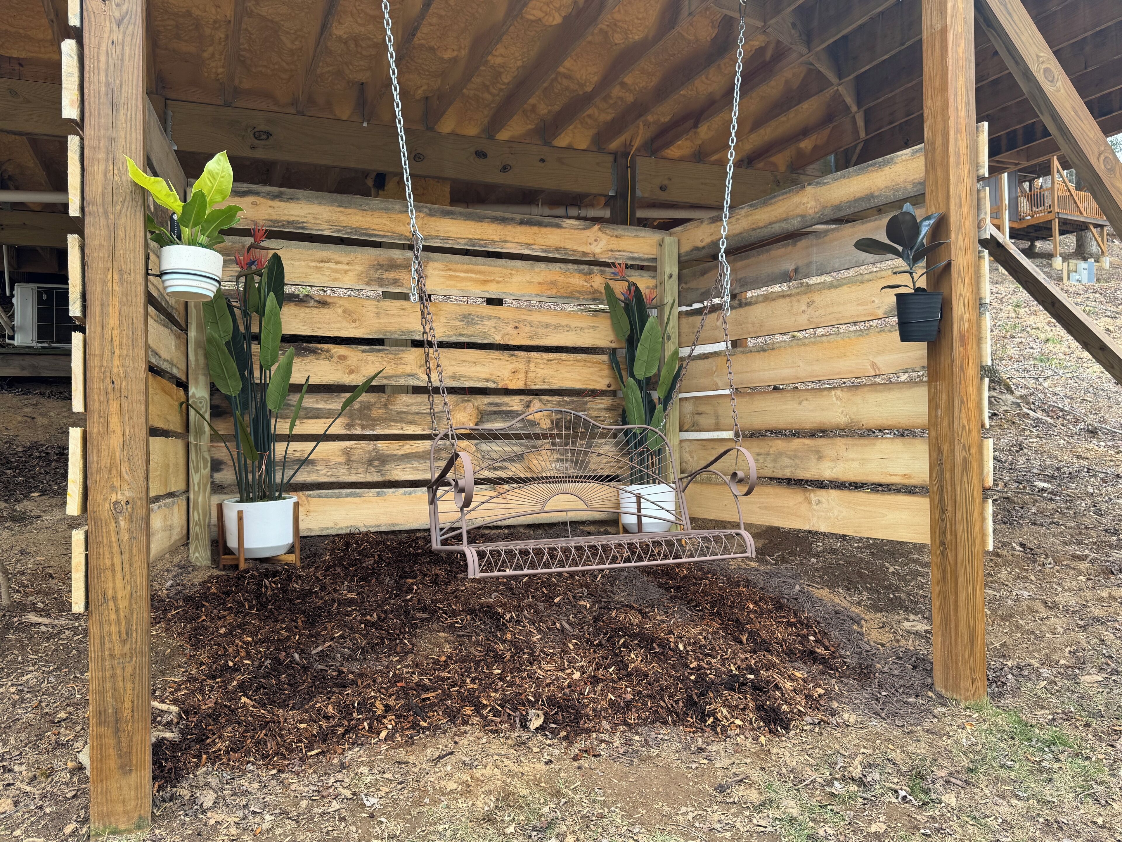 Un fauteuil suspendu en bois, fixé au plafond par des chaînes métalliques, entouré de lattes de bois et de plantes en pot.