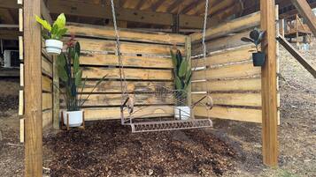 Un fauteuil suspendu en bois, fixé au plafond par des chaînes métalliques, entouré de lattes de bois et de plantes en pot.
