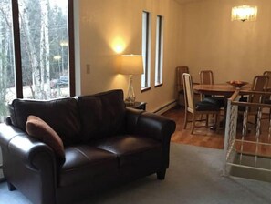 Una sala de estar con un sofá de cuero oscuro, una mesa de comedor con sillas y un ventanal con vista a árboles.
