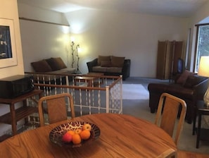 Comedor con una mesa redonda, sillas y un frutero. Se observan dos sofás y un televisor al fondo.