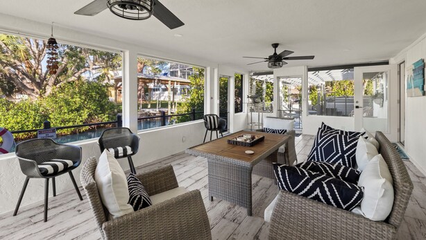 Un espacio exterior techado con muebles de mimbre, ventiladores de techo y grandes ventanales con vistas a una piscina y follaje.