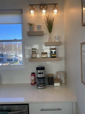A kitchen with a coffee maker, shelves with decorative items, and a window with a view of the neighborhood.