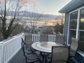 A covered patio with a round table and chairs, a white railing, and a view of a sunset over a residential area.