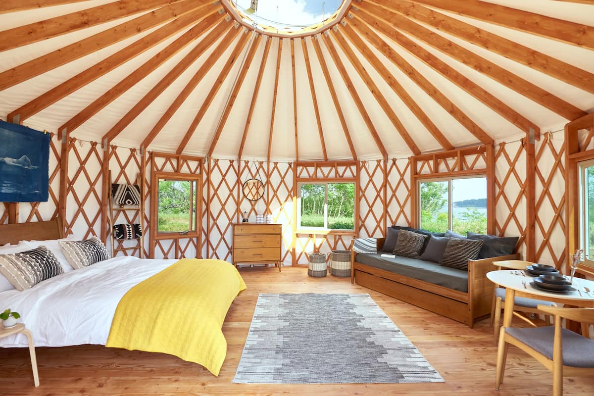 A yurt with a bed, sofa, and dining area, featuring wooden lattice walls and a circular skylight.