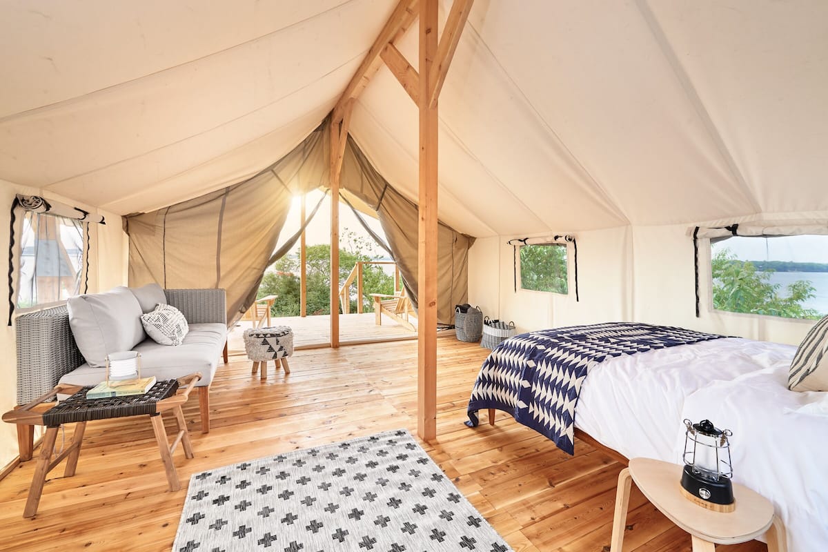 A tented accommodation with a sofa, a bed, a small table, and a view of greenery.