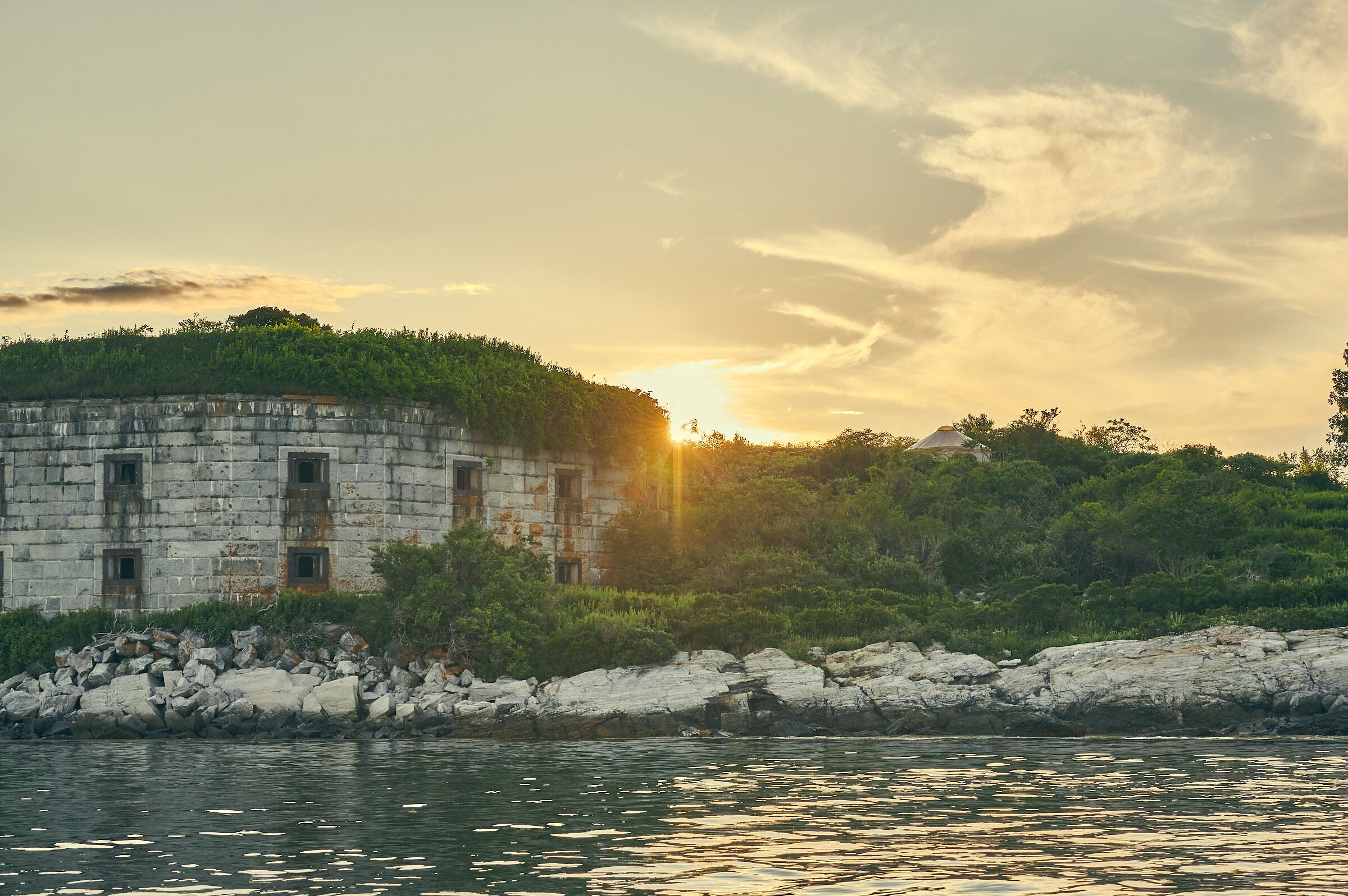 A stone building with multiple windows, surrounded by greenery and a rocky shoreline.