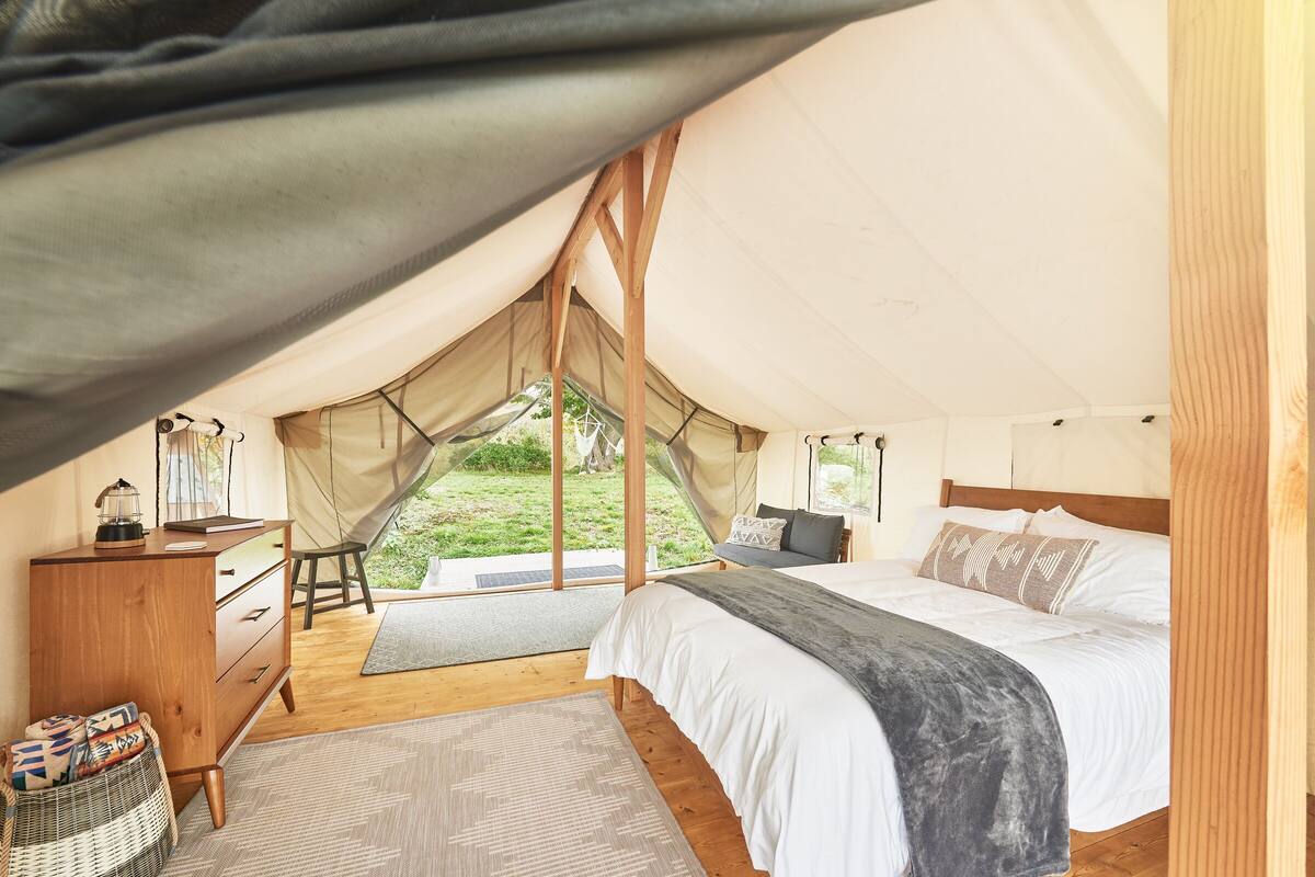 A tented accommodation with a bed, bedside table, and a view of the outdoors.
