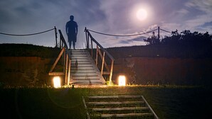 Eine Holzbrücke führt zu einem Gebäude mit beleuchtetem Eingang, unter einem Nachthimmel mit sichtbarischem Mond.
