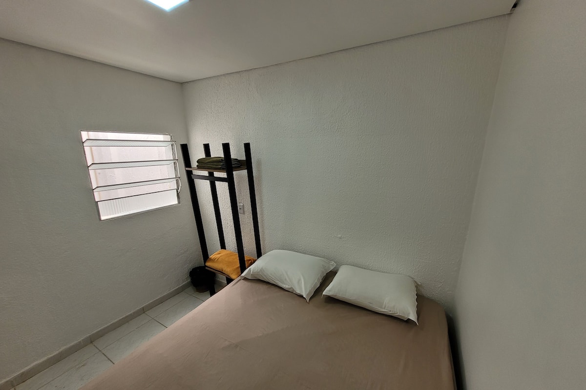 Una habitación pequeña con cama individual, ventana, radiador y un cubo de basura.