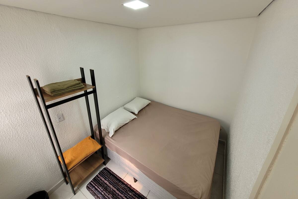 Una habitación pequeña con cama individual y literas en una pared, una mesita de noche y una lámpara en el techo.