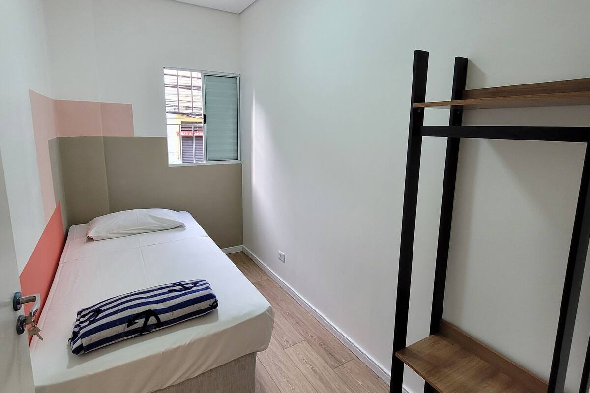 Una habitación pequeña y bien iluminada con una cama individual, una litera de madera, una ventana con persianas y una puerta con picaporte.