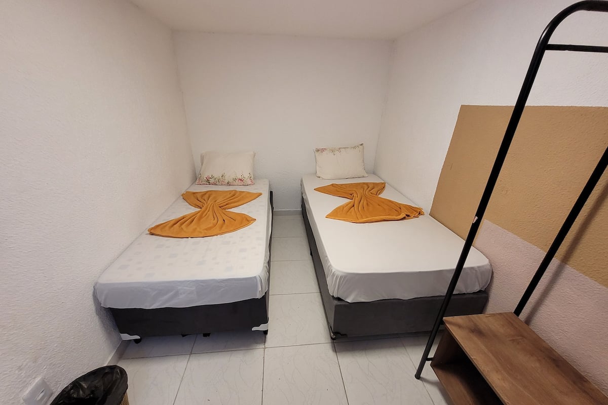 Una habitación pequeña y estrecha con dos camas individuales, cada una con un colchón blanco y una manta marrón. Hay un banco de madera pegado a la pared y una escalera de metal negro.