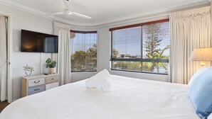 A hotel room with a large bed, a flat-screen TV mounted on the wall, a ceiling fan, and large windows with blinds. - Riverdance Apt 14 (Mooloolaba)