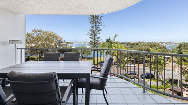 A balcony with a table and chairs, overlooking a coastal area with trees and a view of the sea. - Riverdance Apt 14 (Mooloolaba)