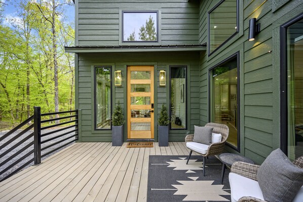 Une terrasse en bois avec des sièges d’extérieur, une porte en bois et une fenêtre donnant sur les arbres.