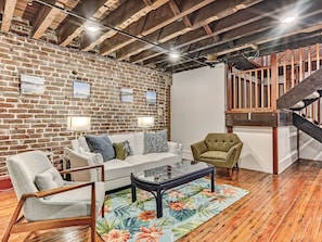 A living room with a brick wall, wooden ceiling beams, a white sofa, a green armchair, and a wooden staircase.