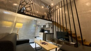 A modern living room with a sofa, a coffee table, and a TV. There is a staircase with a railing and a kitchen area in the background.