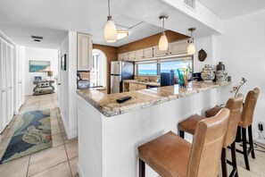 Una cucina moderna con isola in marmo, sgabelli da bar, illuminazione a sospensione e vista sull'oceano.
