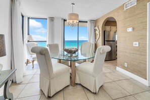 A dining area with a glass table, white upholstered chairs, a chandelier, and a view of the ocean. - Mainsail 131 | Walk to Beach + Balcony + Gulf Views + Seasonal Beach Service (Miramar Beach)