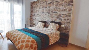Ein Schlafzimmer mit Stehwand, einem Bett mit gemusterten Textilien, einem weißen Stuhl und einem Fenster mit transparenten Vorhängen.