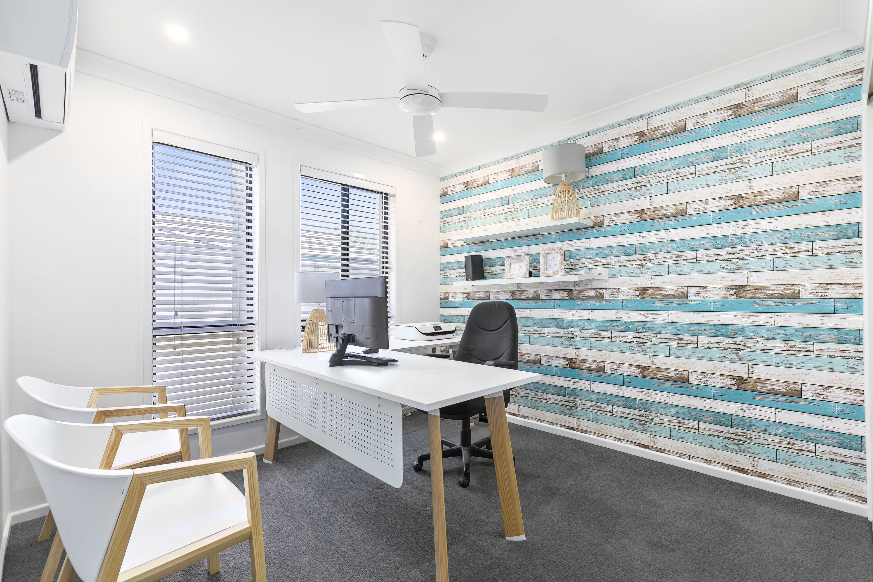 A modern office space with a white desk, black office chair, and two white chairs. There is a ceiling fan, a white air conditioning unit, and a wall with a blue and white distressed wood panel design.