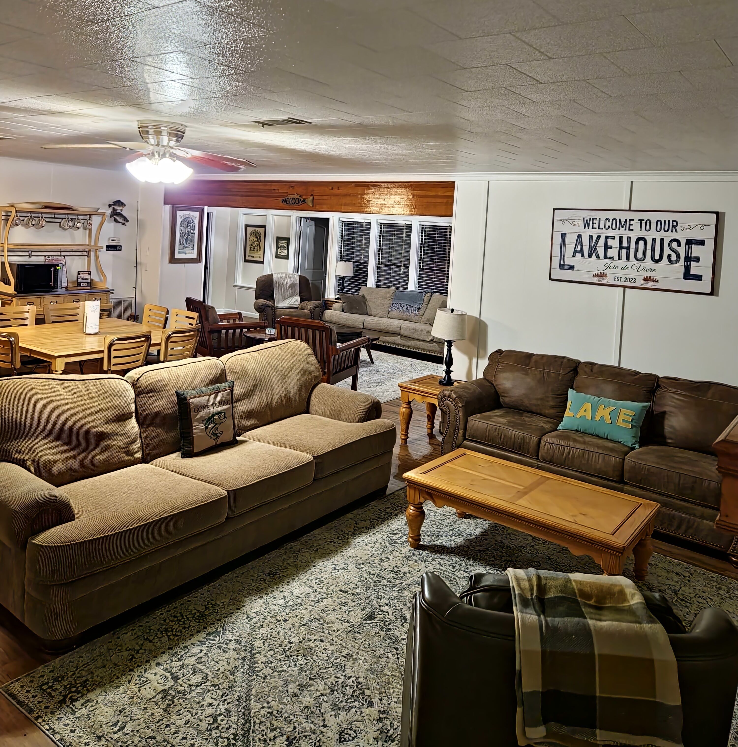 Un salon chaleureux avec une table à manger en bois, deux canapés, une table basse et un panneau indiquant : « Bienvenue dans notre maison au bord du lac. »