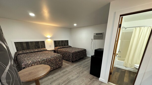 A hotel room with two beds, a small table, a trash bin, and a bathroom visible through an open door.