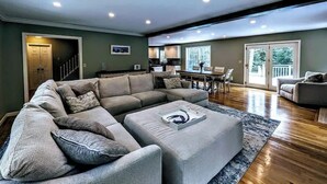 A spacious living room with a sectional sofa, ottoman, and a dining area with a table and chairs. - Charming Bar Harbor Home w/ Fire Pit & Trails (Bar Harbor)