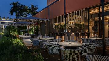 An outdoor seating area with round tables and patterned chairs, surrounded by lush greenery and palm trees.