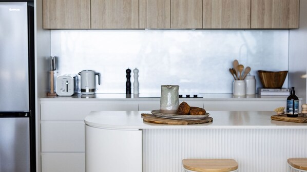 A modern kitchen with white cabinetry, a central island with stools, a coffee maker, and a bottle of olive oil. - 101 on the Beach, Jervis Bay (Huskisson)