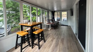 Ein modernes Interieur mit Essbereich, inklusive Holztisch und Barhocker, großen Fenstern mit Blick auf Grünflächen und einer Glasschiebetür zum Außenbereich.