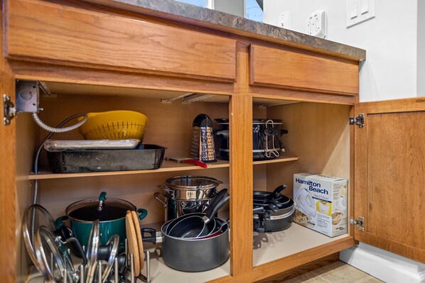 Cocina con armarios de madera, un colador amarillo, varias ollas y sartenes, un rallador y una batidora de mano Hamilton Beach.