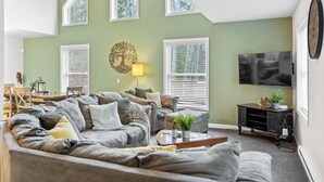 A living room with a large sectional sofa, a flat-screen TV, and a dining area with a table and chairs. - Arrowhead Lake Retreat with large deck and fire pit (Pocono Lake)