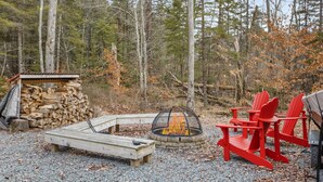 A forested campsite with a fire pit, red Adirondack chairs, and a wooden deck. - Arrowhead Lake Retreat with large deck and fire pit (Pocono Lake)