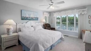 A bedroom with a large bed, a ceiling fan, a nightstand with a lamp, a chair, and a view of a lake through large windows.