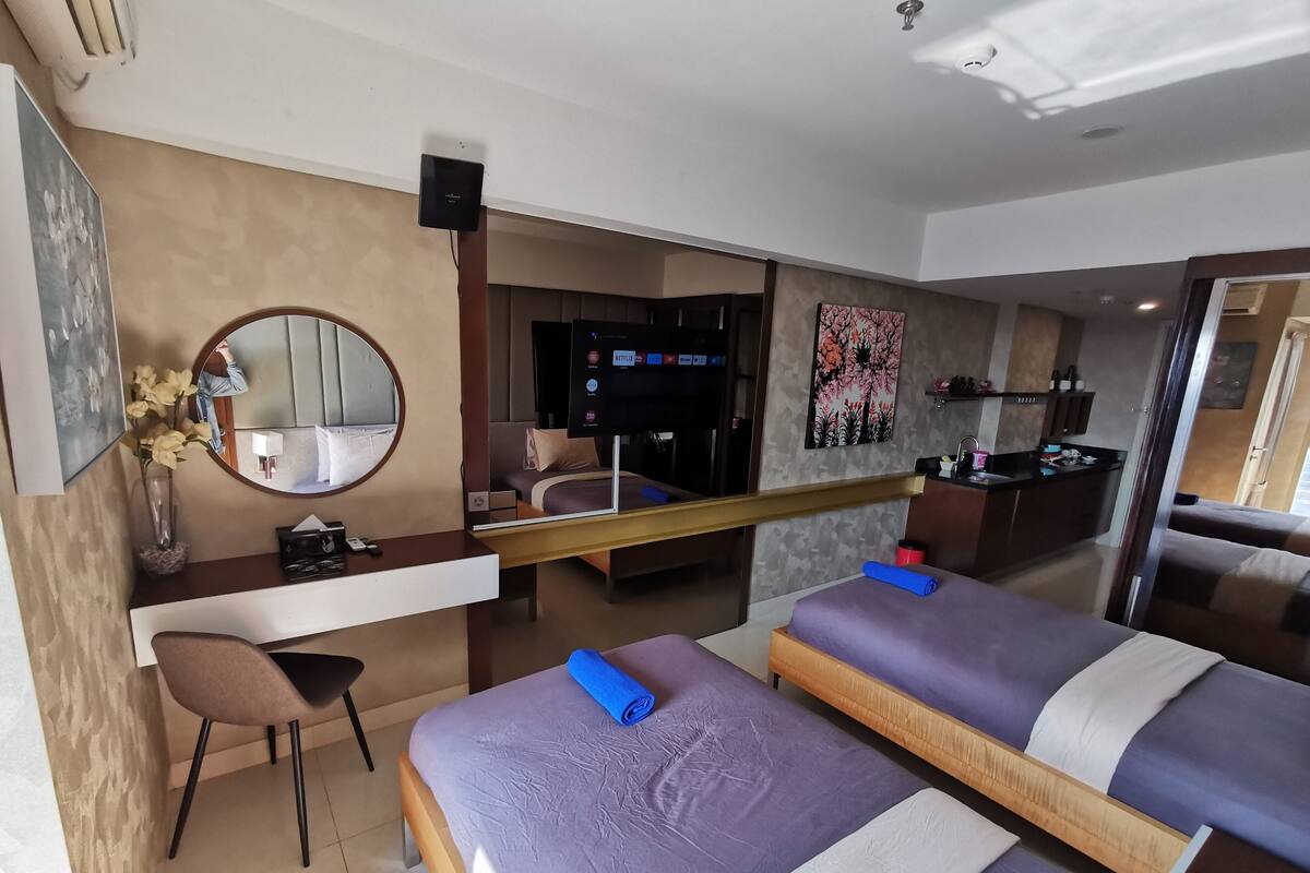 A hotel room with two beds, a desk with a chair, a mirror, a TV, and a kitchenette.