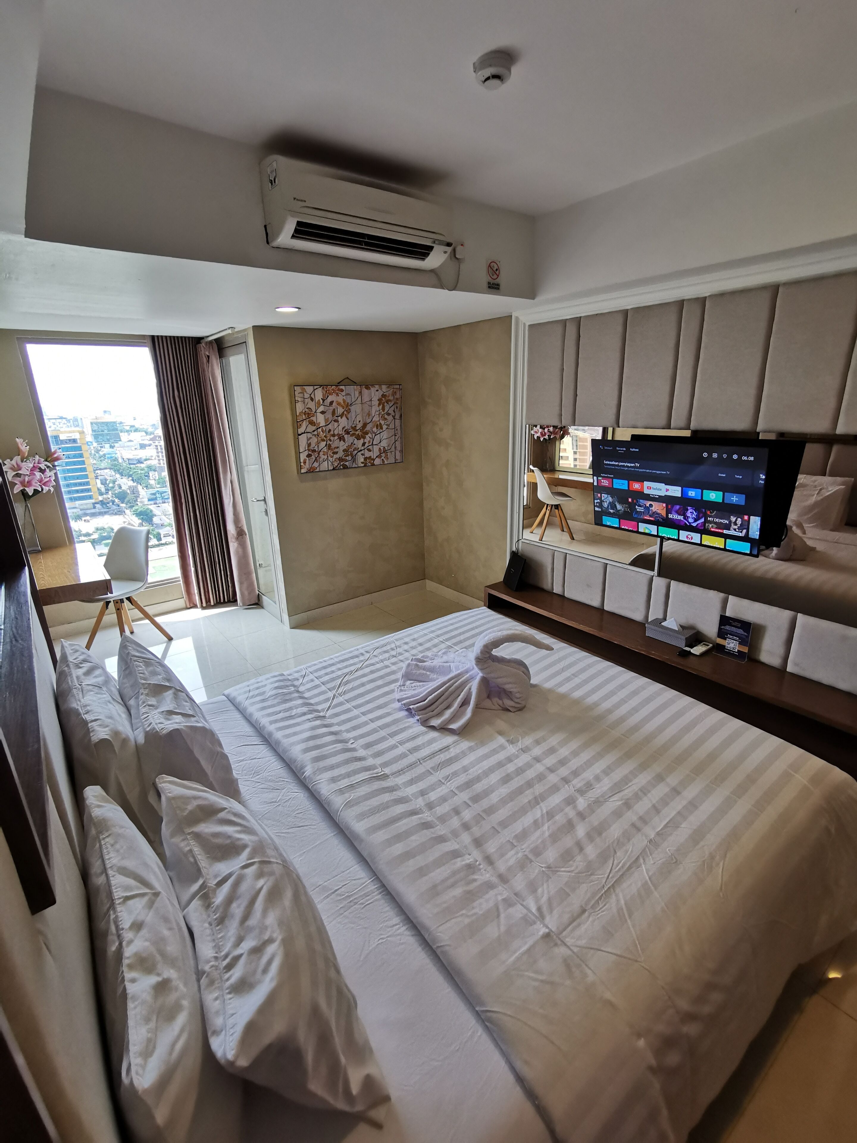 A hotel room with a large bed, a flat-screen TV, an air conditioning unit, a view of the city, and a balcony with flowers.