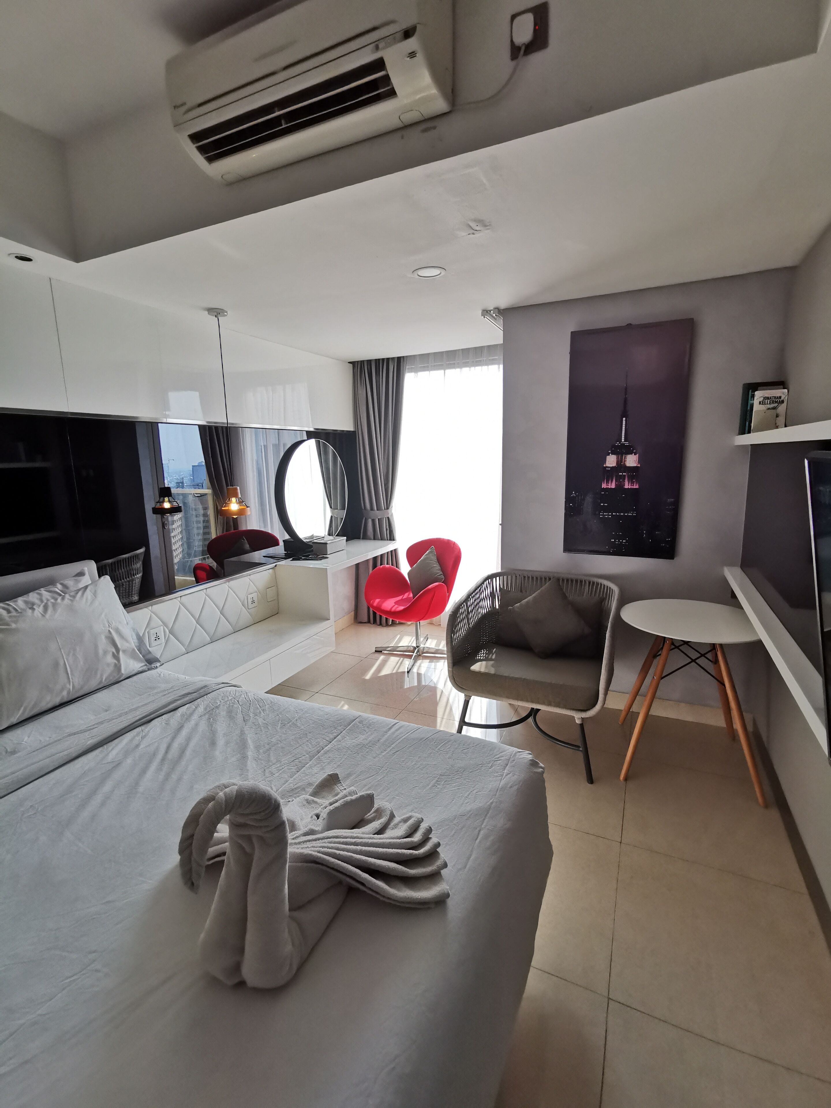A modern hotel room with a large bed, a desk, a red chair, a round mirror, and a cityscape poster.