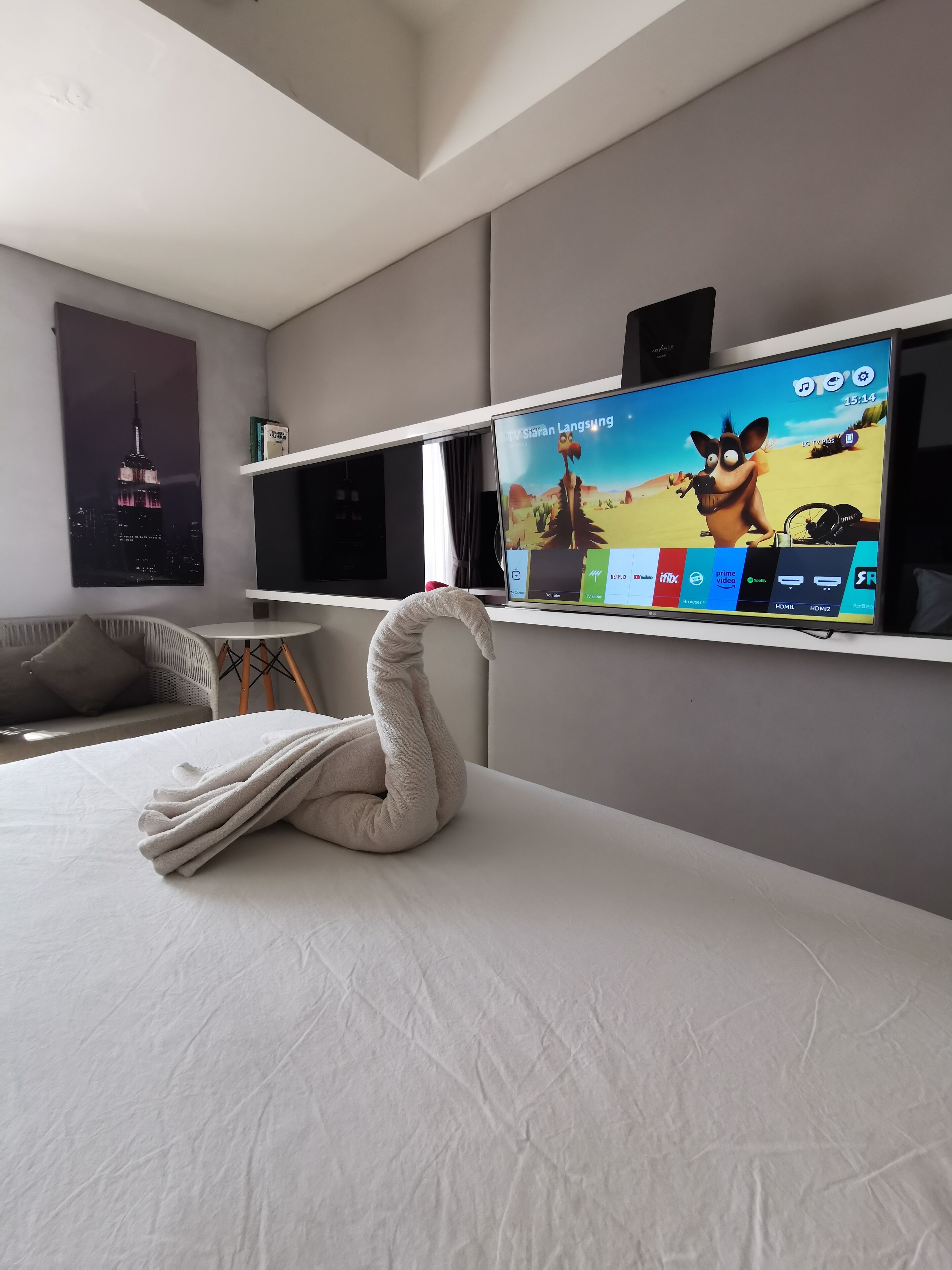 Quarto de hotel moderno com uma grande televisão de tela plana na parede, uma cama bem arrumada com travesseiro em formato de cisne, um sofá cinza e um pôster do Empire State Building.