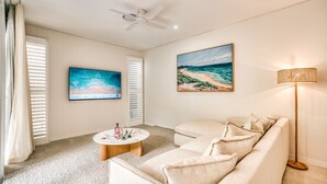 Ein Wohnzimmer mit einem beigefarbenen Sofa, einem runden Couchtisch mit Dekoration, einem Flachbildfernseher und einem Gemälde mit einer Strandansicht.
