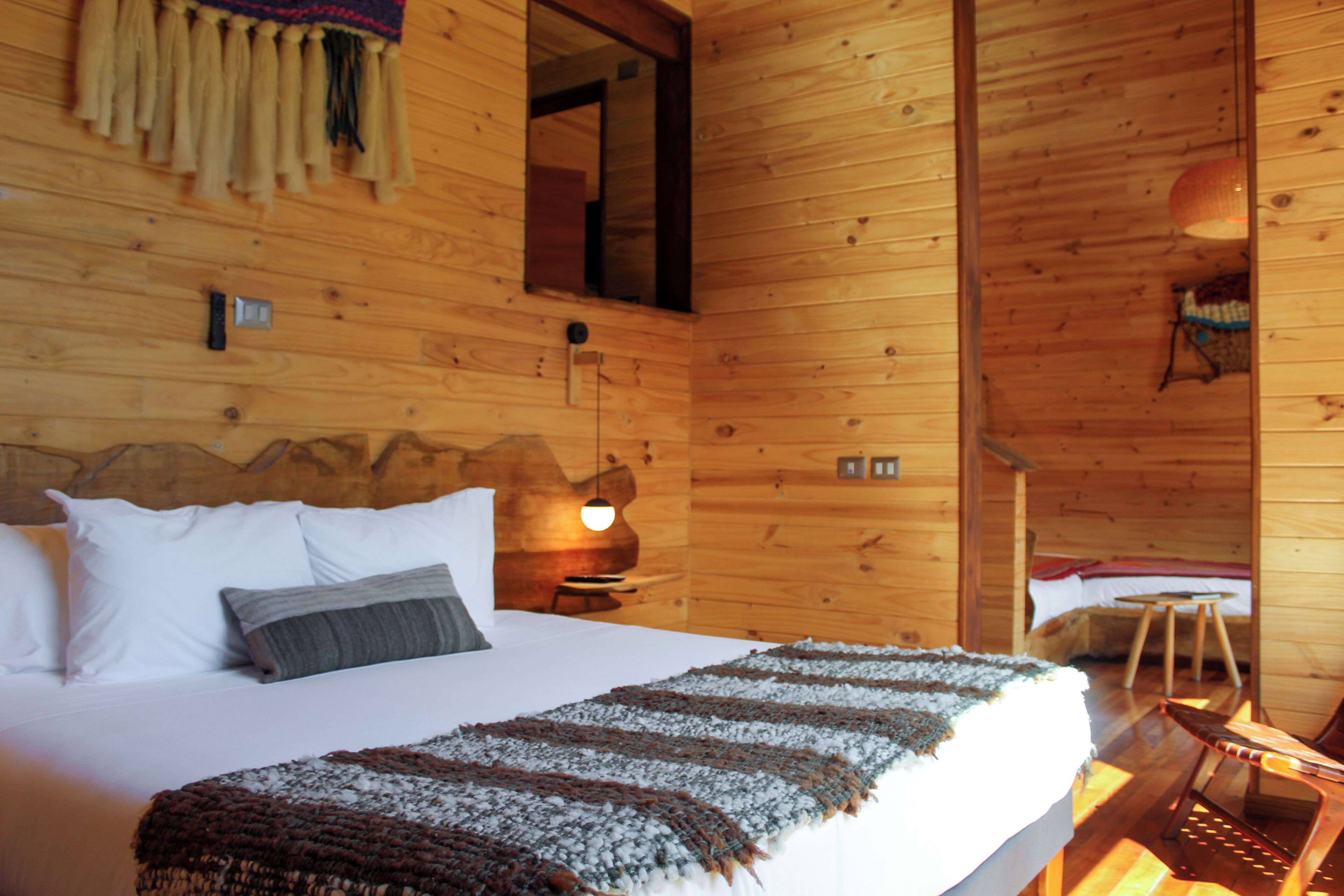 Un dormitorio de cabaña de madera con cama, almohadas, una manta, una lámpara colgante, un espejo y una silla de madera.