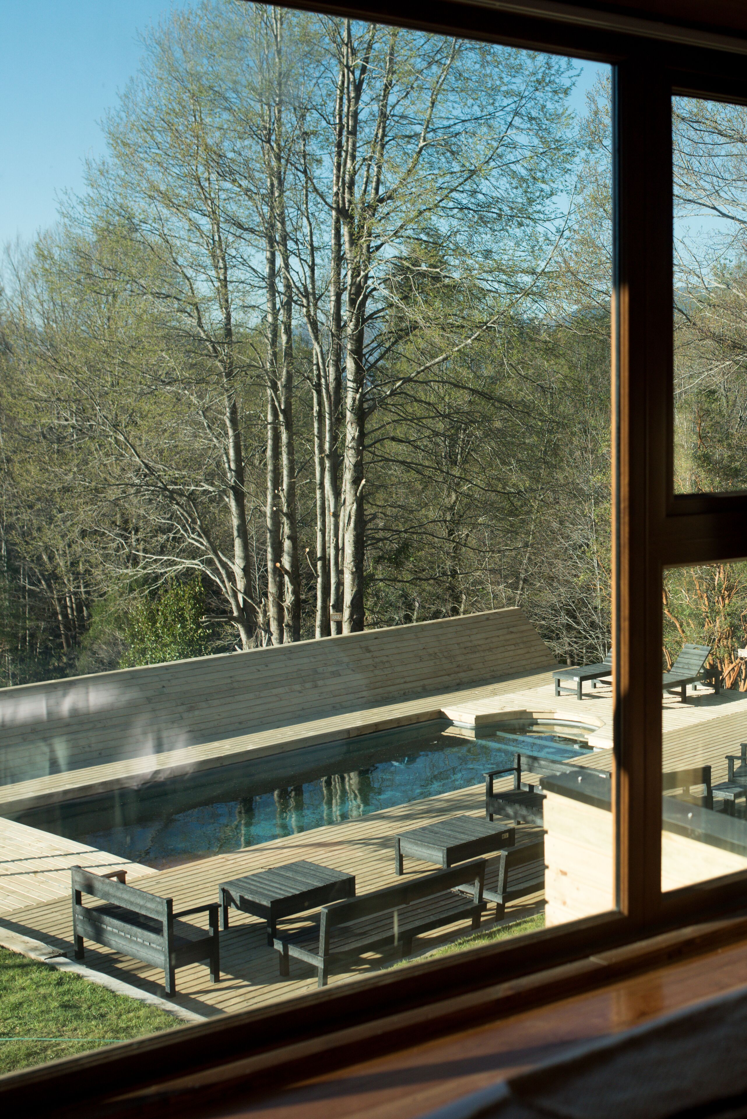 A view of a swimming pool with wooden deck and benches, surrounded by trees.